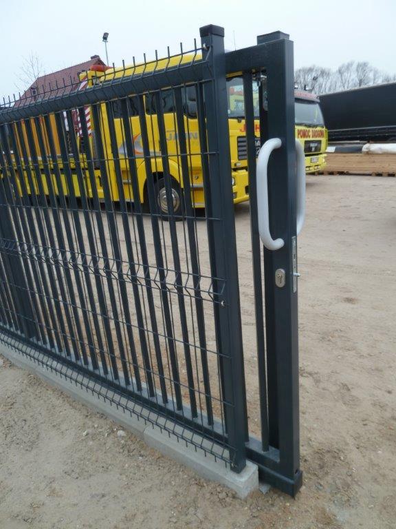 Metalowa furtka panelowa z klamką, osadzona na betonowej podmurówce. W tle widoczny żółty samochód ciężarowy i składowane drewno.