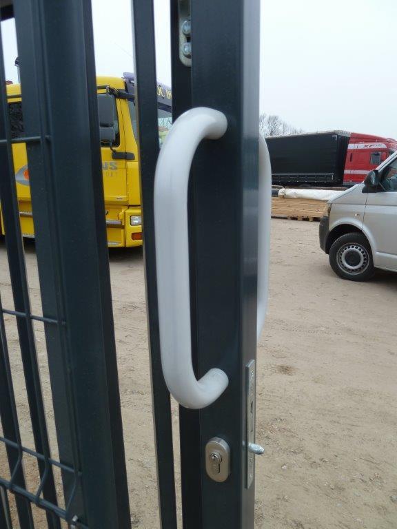 Szczegół zamkniętej, grafitowej bramy z siatki, z białą, ergonomiczną rączką i zamkiem. W tle widoczne pojazdy i materiały budowlane.