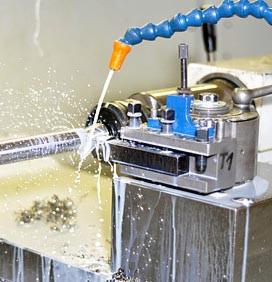 Metalowy pręt obrabiany na tokarce z użyciem chłodziwa, wióry metalowe wokół, widoczna głowica tokarska z narzędziem skrawającym.