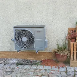 Szara jednostka zewnętrzna pompy ciepła zamontowana na elewacji domu, obok drewnianych skrzynek z kwiatami i doniczkami, widok z zewnątrz.