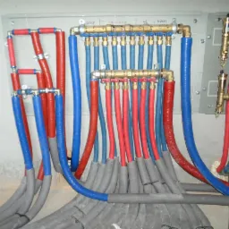 Rozdzielacz ogrzewania podłogowego z rurami w kolorach niebieskim i czerwonym, zabezpieczonymi szarą izolacją. Widoczne oznaczenia pomieszczeń: GOSP, ŁAZIENKA, WC, KUCHNIA.