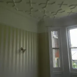 Pokój z paskowaną tapetą w odcieniach beżu, dekoracyjnym sufitem z geometrycznym wzorem i oknem w białej ramie z widokiem na zieleń.