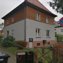 Dwukondygnacyjny dom jednorodzinny z drewnianą elewacją na górnej kondygnacji i szarą na dolnej, z czerwoną dachówką, widoczny z zewnątrz przez metalowe ogrodzenie z numerem posesji.