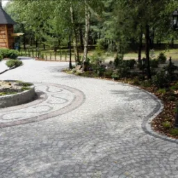 Ogród z ścieżką z kostki granitowej ułożonej w łukowaty wzór, centralnie umieszczone drzewko w okrągłej kamiennej rabacie, w tle drewniana altana i drzewa.