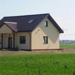 Widok z zewnątrz na beżowy dom jednorodzinny z ciemnobrązowym dachem, otoczony zielonym trawnikiem i polami w oddali.