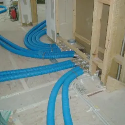 Niebieskie, karbowane przewody wentylacyjne prowadzone pod podłogą w surowym stanie, połączone z aluminiowymi kanałami w ścianie szkieletowej, widoczne narzędzia i resztki materiałów budowlanych.