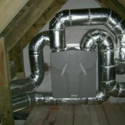 Szara centrala wentylacyjna z izolowanymi, srebrnymi przewodami w przestrzeni poddasza z drewnianą konstrukcją dachu.