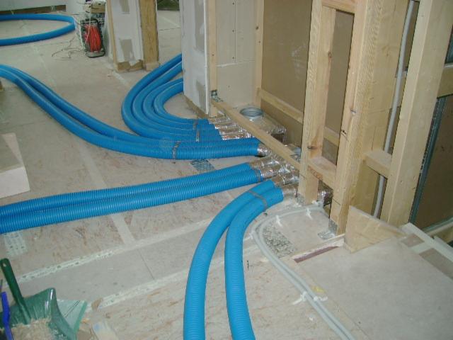 Niebieskie, karbowane przewody wentylacyjne prowadzone pod podłogą w surowym stanie, połączone z aluminiowymi kanałami w ścianie szkieletowej, widoczne narzędzia i resztki materiałów budowlanych.