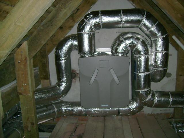 Szara centrala wentylacyjna z izolowanymi, srebrnymi przewodami w przestrzeni poddasza z drewnianą konstrukcją dachu.