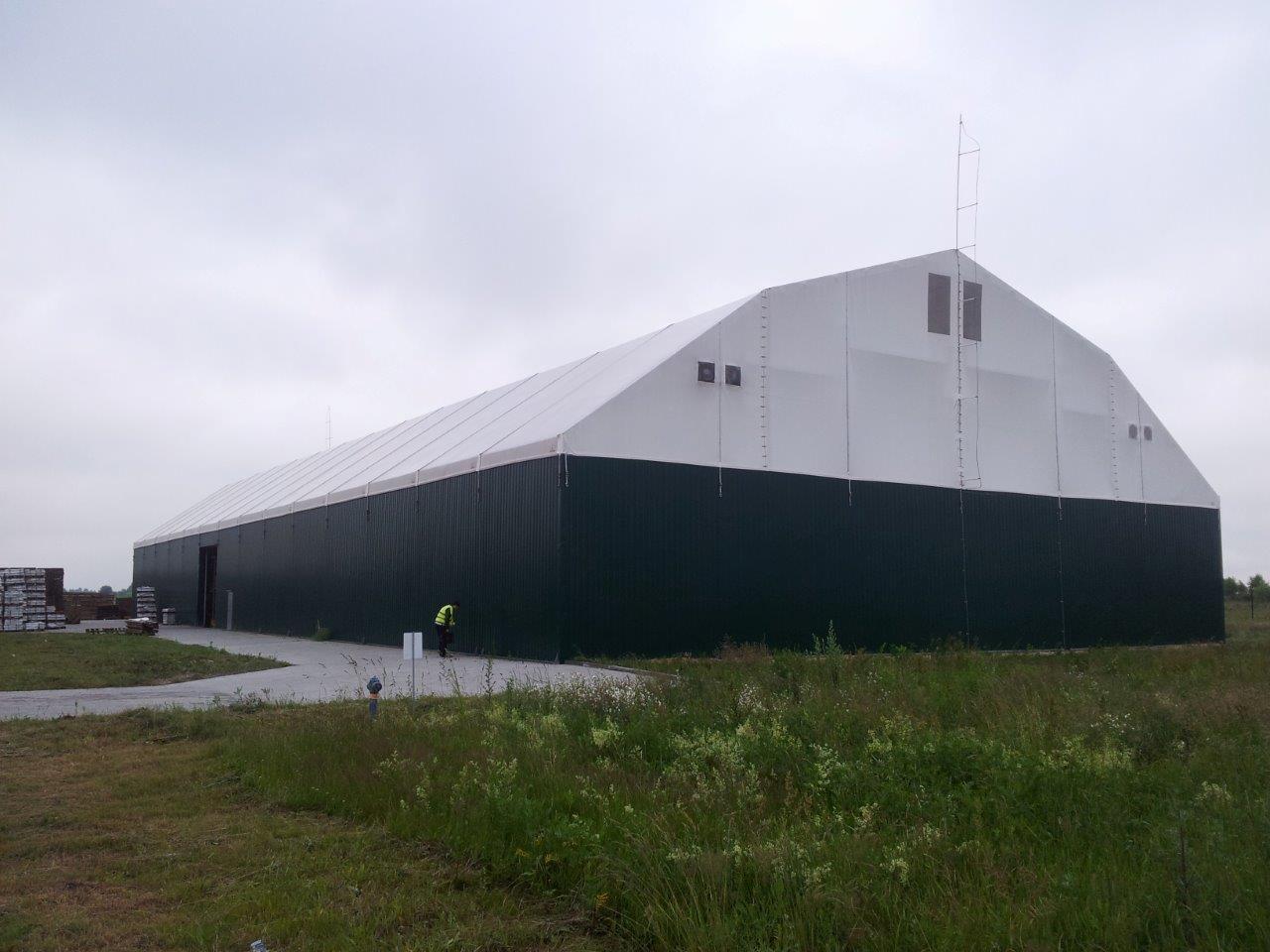 Duża hala namiotowa z zielonymi ścianami i białym dachem, widoczna z zewnątrz, z pracownikiem w żółtej kamizelce przed wejściem, na tle pochmurnego nieba.
