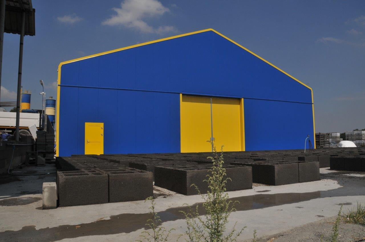 Hala magazynowa z niebieskimi ścianami i żółtymi drzwiami przesuwnymi, przed którą ułożone są betonowe bloczki.