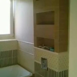 Nowoczesna aranżacja łazienki w Sochaczewie: beżowe płytki imitujące drewno na ścianie z wnękami i dekoracyjne pasy. Umywalka częściowo widoczna, jasne wnętrze.