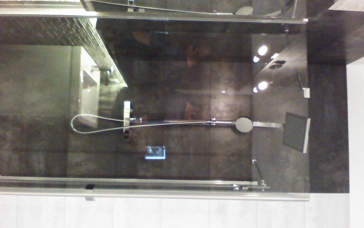 Nowoczesna kabina prysznicowa z deszczownicą i szklanymi drzwiami, wykończona ciemnymi płytkami. Widok z góry, z odbiciem osoby w szkle.