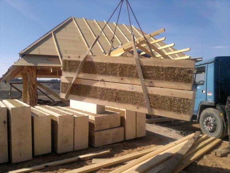 Bloki ze słomy w drewnianej ramie podnoszone dźwigiem na placu budowy. Widoczny szkielet dachu oraz paleta bloków. W tle niebieska ciężarówka.