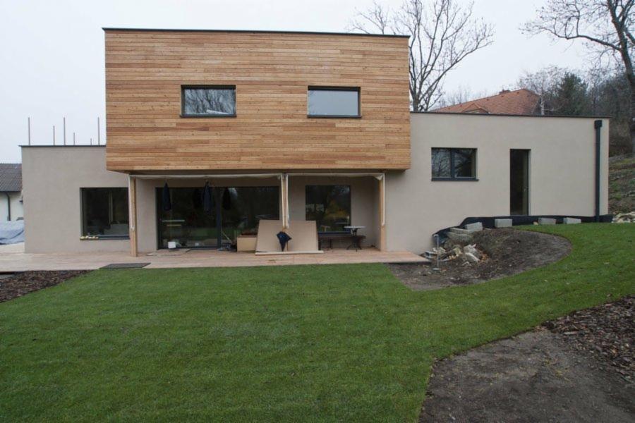 Nowoczesny dom jednorodzinny w trakcie budowy, z elewacją częściowo wykończoną drewnem, widoczne elementy konstrukcyjne tarasu i świeżo założony trawnik.