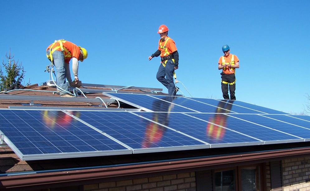 Montaż paneli fotowoltaicznych na dachu domu jednorodzinnego; trzech monterów w kaskach i uprzężach zabezpieczających pracuje na dachu w słoneczny dzień.