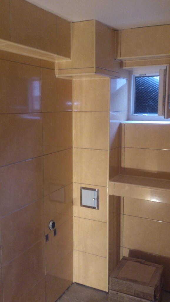 Wykończona łazienka z beżowymi, błyszczącymi płytkami ceramicznymi, zabudową nad oknem i ukrytym zaworem wodnym, widoczne kartony na podłodze.