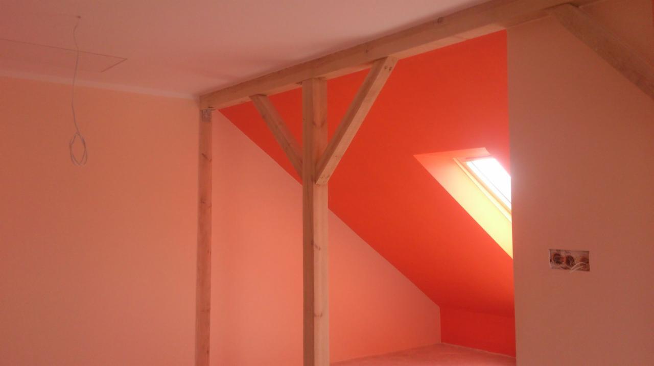 Poddasze w trakcie remontu z widoczną drewnianą konstrukcją nośną, pomalowane ściany w dwóch odcieniach pomarańczowego, okno dachowe i wystające kable elektryczne.