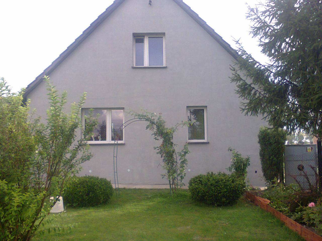 Elewacja domu jednorodzinnego pomalowana na szaro, z widocznymi oknami, zielenią w ogrodzie i metalową pergolą.