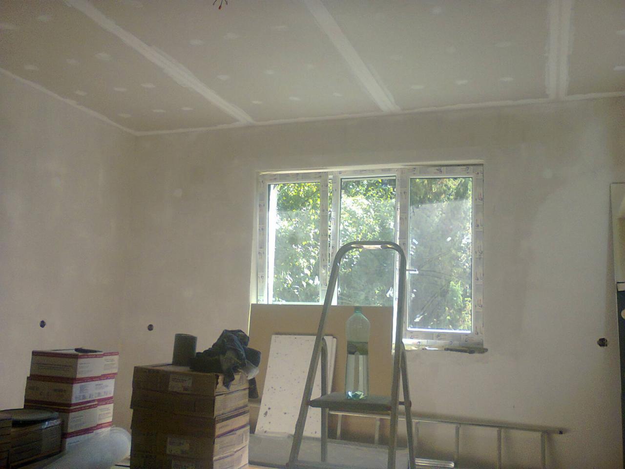 Pokój w trakcie remontu: ściany i sufit po szpachlowaniu, widoczne łączenia płyt karton-gips, okno z widokiem na zieleń, drabina aluminiowa z butelką wody na stopniu, kartony z materiałami...