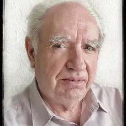 Portret starszego mężczyzny z siwymi włosami i niebieskimi oczami, ubrany w koszulę w paski, na tle białej ściany.