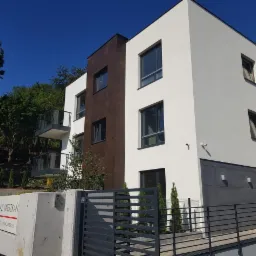 Nowoczesny, trzypiętrowy budynek z białymi ścianami i brązowymi panelami, z balkonami z szklanymi balustradami, widok z poziomu ulicy przez czarne metalowe ogrodzenie.