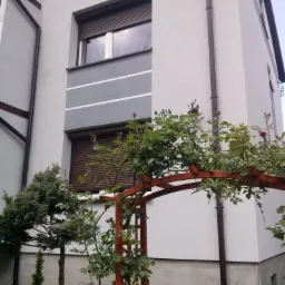 Szary dom z brązowymi roletami zewnętrznymi i drewnianą pergolą z różami, widok z dołu.