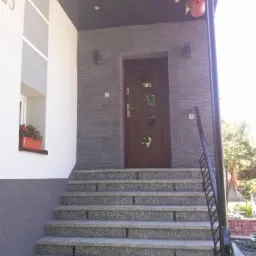 Wejście do domu z drewnianymi drzwiami zewnętrznymi z przeszkleniami o geometrycznym kształcie, wyłożone kamieniem elewacyjnym w odcieniach szarości, z granitowymi schodami wejściowymi i kwiatami...