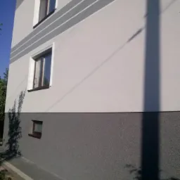 Fragment elewacji dwukondygnacyjnego domu z oknami, pomalowanej na dwa odcienie szarości z dekoracyjnymi pasami, cień słupa energetycznego na ścianie.