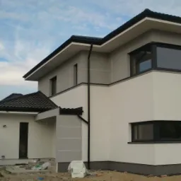 Nowoczesny dom dwukondygnacyjny z szarą i białą elewacją, czarnymi oknami PCV i rynnami, widoczne materiały budowlane na niezagospodarowanym terenie.