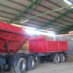 Czerwona ciężarówka z otwartą skrzynią ładunkową, wysypująca żwir na ziemię wewnątrz zadaszonej hali z ceglanymi ścianami.