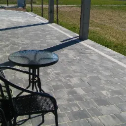 Szara kostka brukowa ułożona na tarasie z widocznym okrągłym stolikiem i krzesłami ogrodowymi, w tle łąka i budynek.