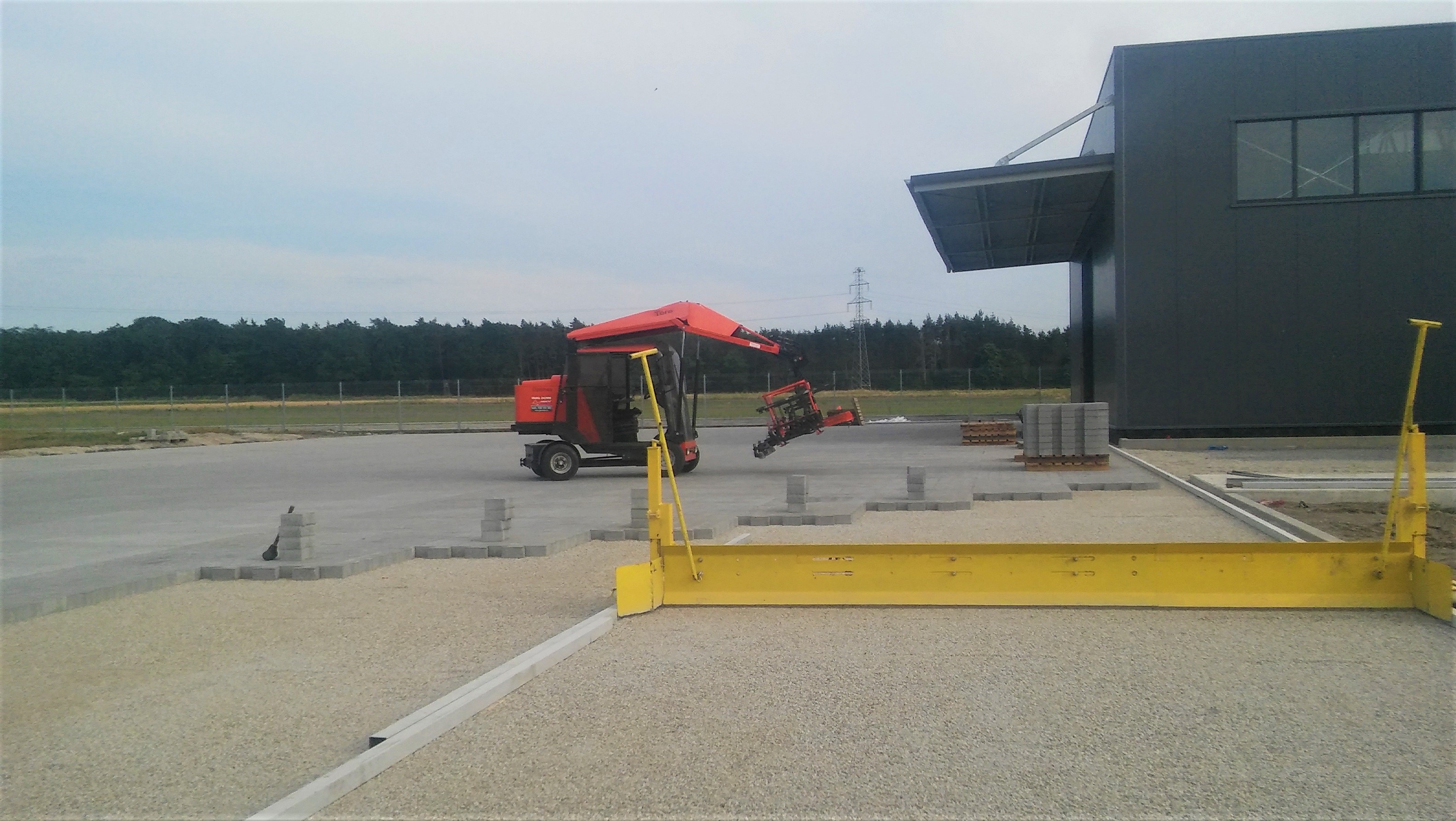 Specjalistyczna maszyna układa kostkę brukową na przygotowanym podłożu w miejscowości Murów, widoczna żółta listwa wyrównująca, w tle budynek przemysłowy.