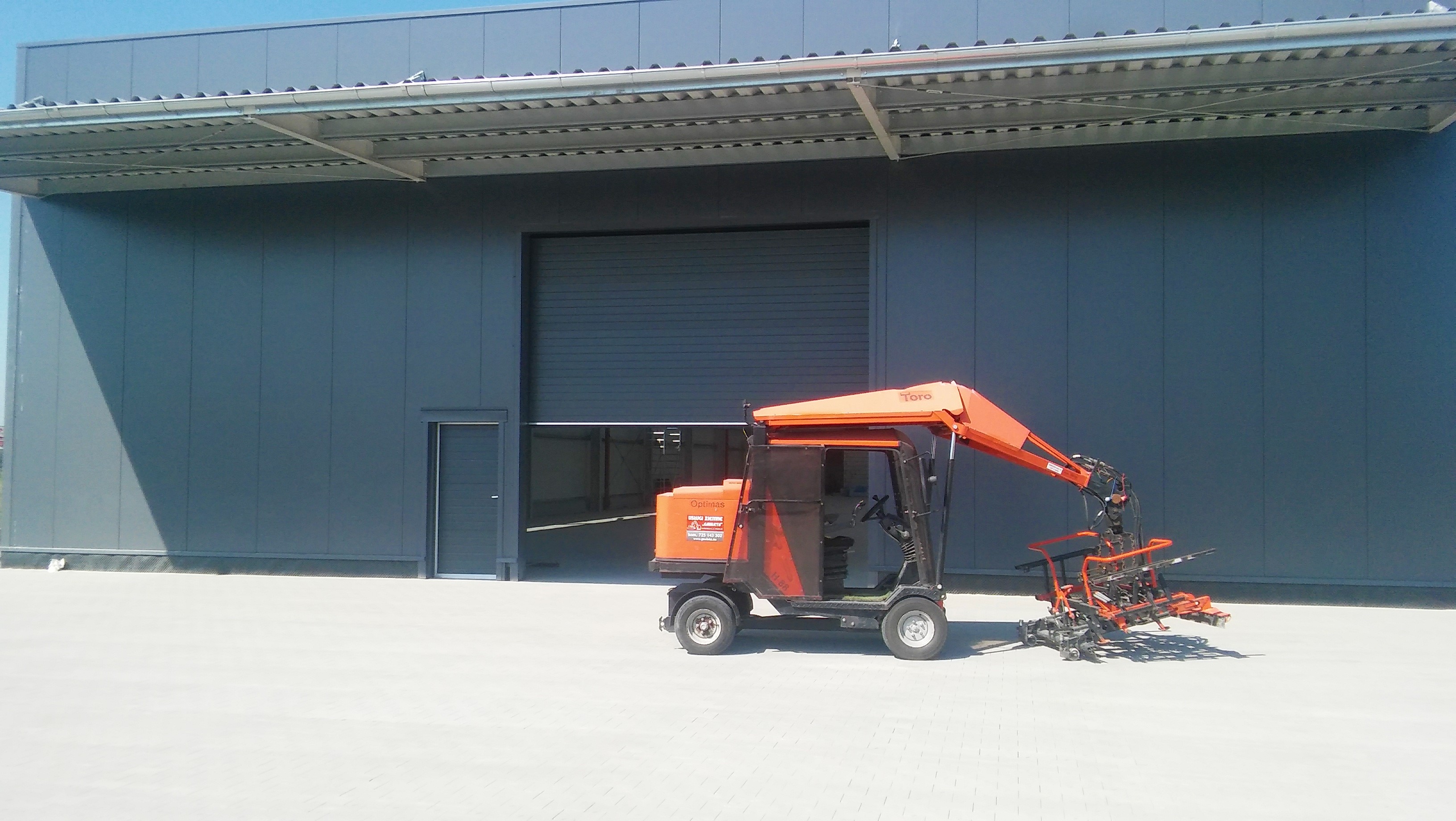Pomarańczowy manipulator próżniowy Toro Optimas FL1.6B na tle szarego, nowoczesnego budynku magazynowego z bramą rolowaną i drzwiami wejściowymi.