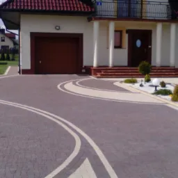 Kaskadowy podjazd z kostki brukowej w odcieniach szarości i beżu, z białymi liniami i obwódkami, prowadzący do domu z garażem i schodami wejściowymi.