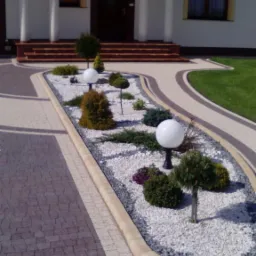 Kaskadowe schody z jasną elewacją budynku. Ogródek z białym żwirem, kulistymi lampami i ozdobnymi krzewami. Brązowa kostka brukowa i jasne obrzeża.