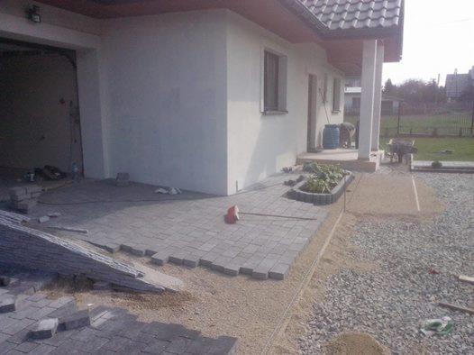 Fragment układania szarej kostki brukowej na podwórku domu jednorodzinnego. Część kostki już ułożona, widoczne narzędzia i materiały budowlane.