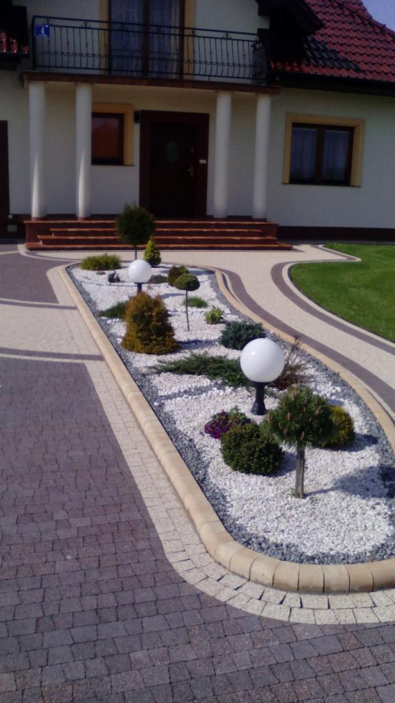 Kaskadowe schody z jasną elewacją budynku. Ogródek z białym żwirem, kulistymi lampami i ozdobnymi krzewami. Brązowa kostka brukowa i jasne obrzeża.