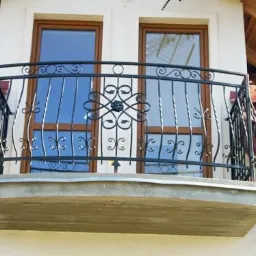 Kuty balkon z ozdobnymi elementami na tle okien i dachu z czerwonej dachówki. Balkon wykonany w stylu klasycznym, z widoczną betonową płytą.