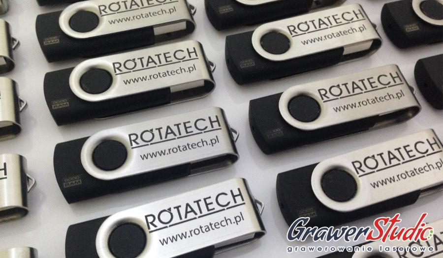 Seria czarnych pendrive'ów z obrotową, srebrną obudową, na której wygrawerowano logo 'ROTATECH' i adres strony internetowej, ułożonych na białym tle, z widocznym w prawym dolnym rogu logo firmy...