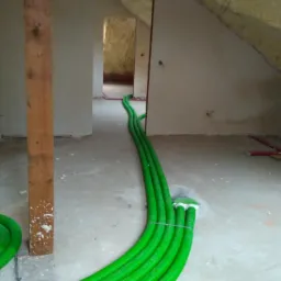 Zielone rury wentylacyjne prowadzone po podłodze w surowym wnętrzu poddasza, widoczne przejście do kolejnego pomieszczenia.