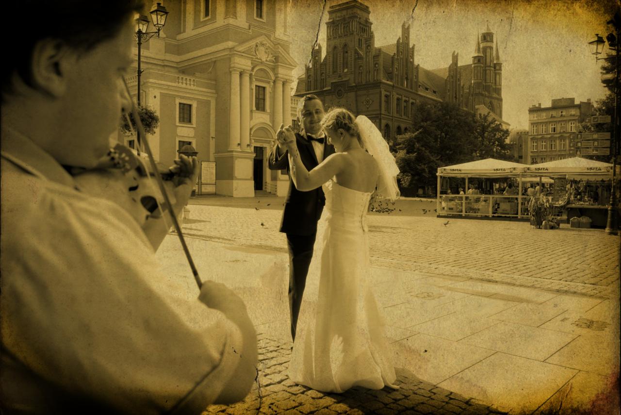 Para młoda tańczy na brukowanym placu w otoczeniu zabytkowej architektury, przygrywa im skrzypek. Stylizacja retro nadaje zdjęciu unikalny charakter.