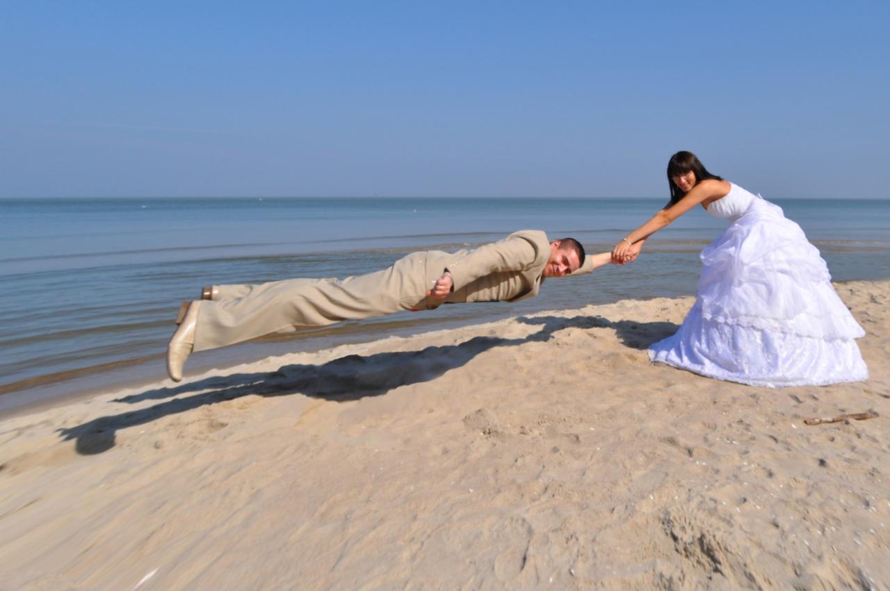 Panna młoda w białej sukni trzyma za rękę leżącego poziomo na piasku pana młodego w beżowym garniturze, morze i błękitne niebo w tle. Nietypowe ujęcie.
