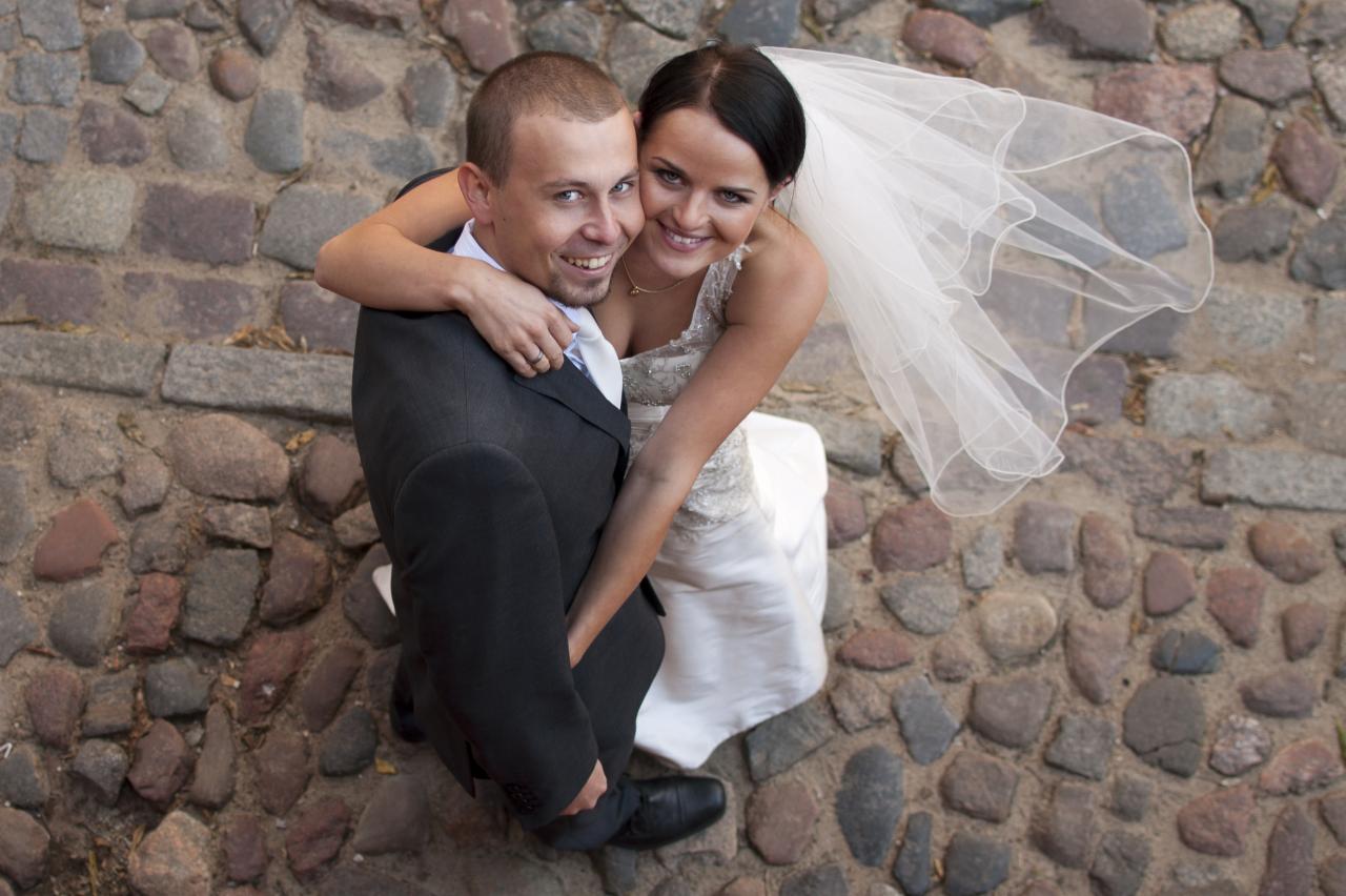 Uśmiechnięta para młoda w objęciach na brukowanej uliczce w Wąbrzeźnie. Panna młoda w welonie, pan młody w garniturze. Ujęcie z góry, podkreślające intymność.