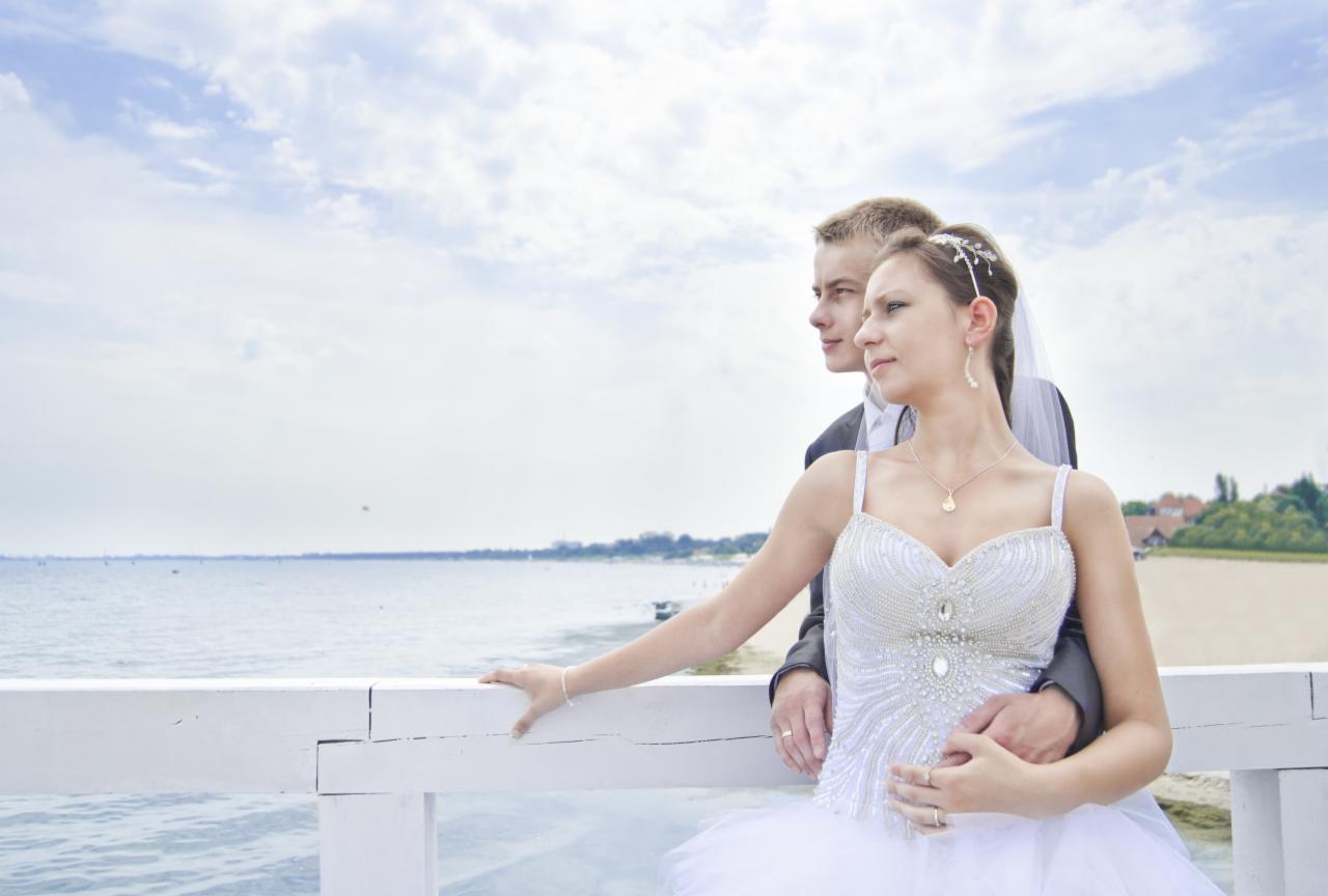 Para młoda na molo, bliskość i zaduma. Panna młoda w sukni z kryształkami, delikatny welon, on w eleganckim garniturze. Tło: morze i błękitne niebo.