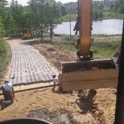 Żółta koparka z łyżką pełną ziemi w trakcie prac ziemnych przy układaniu ażurowej kostki brukowej na drodze w pobliżu jeziora, widok z kabiny operatora.