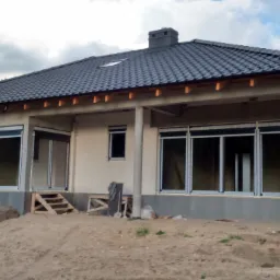 Niedokończony dom jednorodzinny z szarymi roletami zewnętrznymi, czarną dachówką i widocznymi fundamentami w piaszczystym terenie.