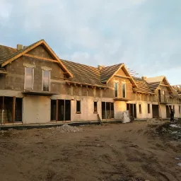 Wznoszony budynek w stanie surowym otwartym, z widoczną konstrukcją dachu i wstawionymi oknami, na placu budowy z rozsypanymi materiałami.