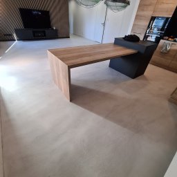 BB DESIGN - Jasnoszara, gładka posadzka z wykończeniem imitującym beton, widoczny fragment drewnianego blatu i zabudowy kuchennej, oraz dekoracyjna ściana z drewnianych listewek.