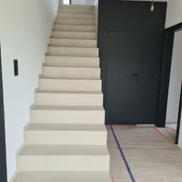 Nowoczesne, minimalistyczne schody o jasnobeżowym odcieniu, prowadzące na wyższą kondygnację, z czarną ścianą i drzwiami w tle, zabezpieczone folią malarską na podłodze.